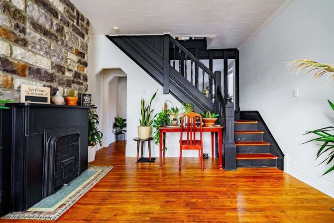 Cozy modern-vintage foyer with glossy hardwood floors, stone accent wall, black vintage fireplace and painted black staircase, red wooden desk and chair topped with potted houseplants