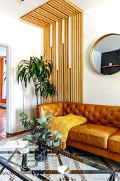Cozy modern living room with caramel tufted leather corner sofa, mustard yellow throw, vertical wooden slat accent wall reaching the ceiling, round gold mirror, tall potted plant, glass coffee table with eucalyptus vase and patterned rug.