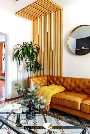 Cozy modern living room with caramel tufted leather corner sofa, mustard yellow throw, vertical wooden slat accent wall reaching the ceiling, round gold mirror, tall potted plant, glass coffee table with eucalyptus vase and patterned rug.