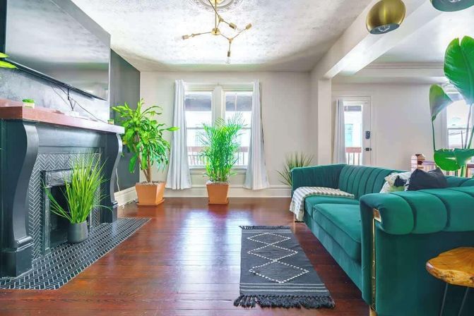 Sunlit modern apartment living room with emerald green velvet sectional, black decorative fireplace, dark hardwood floors, potted indoor plants, large windows with white curtains and brass chandelier.