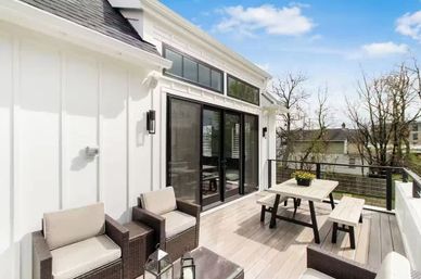 Sunlit modern rooftop deck with white board-and-batten siding and black-framed sliding glass doors, wicker lounge chairs with beige cushions, a wooden picnic table with bench and planter, and neighborhood tree views.