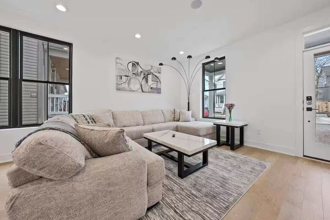 Bright modern living room with L-shaped beige sectional, marble-top coffee table, black-framed windows, arching floor lamp and light hardwood floors.