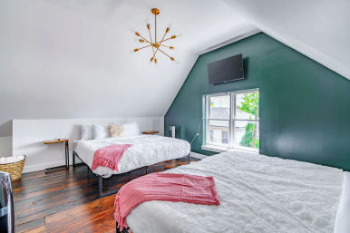Cozy attic bedroom with two beds, fluffy white bedding and pink throws, dark wood floors, green accent wall with mounted TV, sloped ceiling and brass chandelier, window view of neighborhood