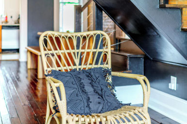 Light rattan armchair with a dark textured macramé cushion on glossy hardwood floors beside a black staircase in a cozy modern loft interior
