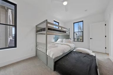 Bright modern bedroom with gray bunk bed and pull-out trundle, white bedding, carpeted floor, and large black-framed windows.