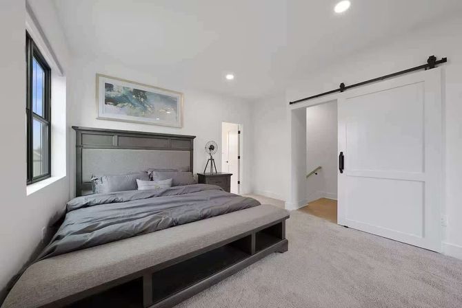 Bright contemporary master bedroom with gray bedding and dark wood headboard, upholstered storage bench at the foot, black-framed window, nightstand with tripod fan, neutral carpet, and white sliding barn door.