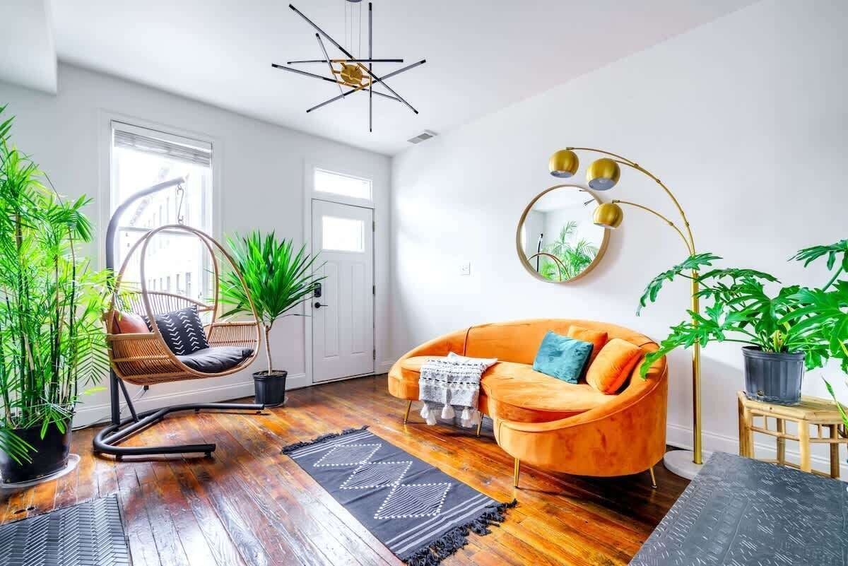 Bright sunlit living room with a curved orange velvet sofa, teal accent pillow, gold arc floor lamp and round mirror, rattan hanging chair by a white front door, dark hardwood floors with black geometric rugs, and lush potted indoor plants for a cozy boho-modern vibe.