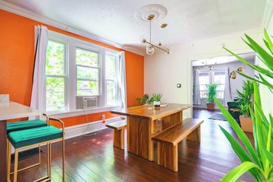 Bright modern dining room with bold orange accent wall, triple windows with a window air conditioner, solid wood bench-style dining table, green bar stools, hanging globe pendant, potted plants, and glossy hardwood floors.
