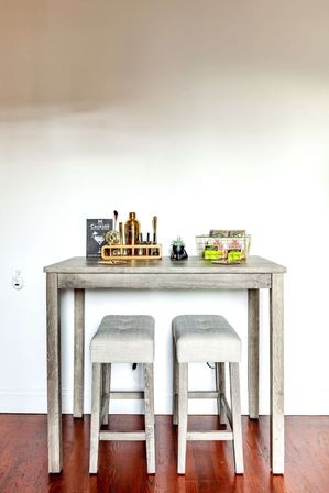 Cozy modern farmhouse mini bar: gray wooden high table with two upholstered stools on hardwood floor, gold cocktail shaker set, small succulent and snack basket against a clean white wall