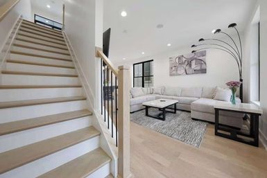 Bright, airy modern living room with light wood staircase, open-concept layout, light hardwood floors, neutral L-shaped sectional, marble-top coffee and side tables, textured rug, arched multi-head floor lamp and abstract wall art