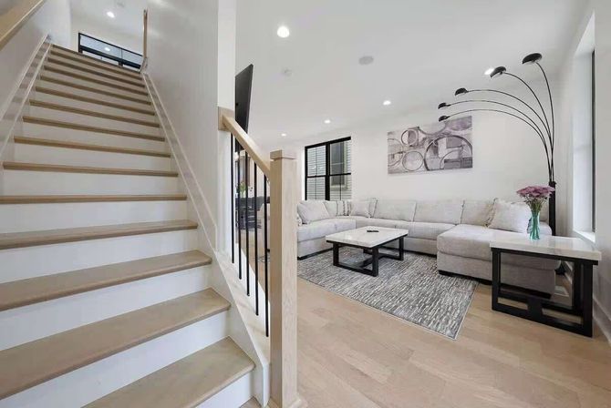 Bright, airy modern living room with light wood staircase, open-concept layout, light hardwood floors, neutral L-shaped sectional, marble-top coffee and side tables, textured rug, arched multi-head floor lamp and abstract wall art