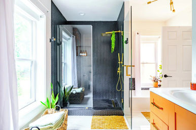 Sunlit modern home bathroom with large window, glass-enclosed walk-in shower lined in black herringbone tile, gold/brass shower fixtures, orange vanity, woven mats and potted plants.
