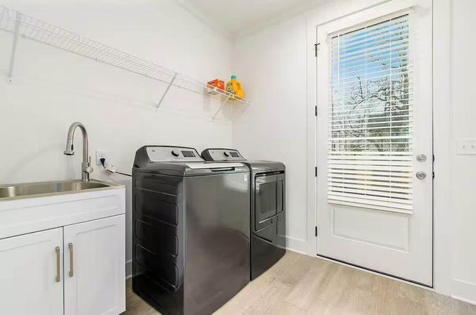 Bright modern residential laundry room with charcoal washer and dryer, white sink cabinet, wire shelf with detergent, and a glass exterior door with blinds letting in natural light.