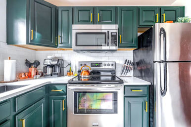 Bright modern L-shaped kitchen with deep green cabinets and brass pulls, white subway tile backsplash, stainless-steel fridge, microwave and oven, copper kettle on a glass-top stove, coffee maker and knife block on white countertops.