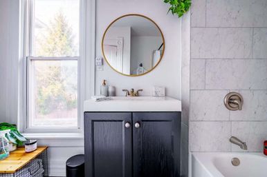 Bright modern bathroom with black vanity and white sink, brass faucet beneath round gold mirror, marble-tiled tub and shower, large window with natural light, wooden bench with plants and toiletries.