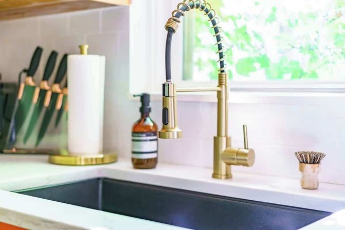 Modern kitchen sink with matte black undermount basin, brushed gold spring pull-down faucet, white quartz countertop, knife block, paper towel holder, amber soap dispenser and small dish brush by a sunny window