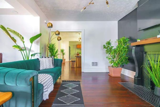 Bright modern living room with emerald velvet sofa, patterned throw and geometric rug, tall indoor plants, brass multi-head floor lamp, dark fireplace mantel, warm hardwood floors and open doorway to kitchen.