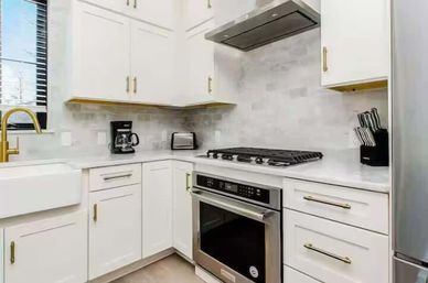 Bright modern L-shaped white kitchen with brass hardware, farmhouse sink, stainless-steel oven and gas cooktop, stone-look backsplash and countertop appliances.