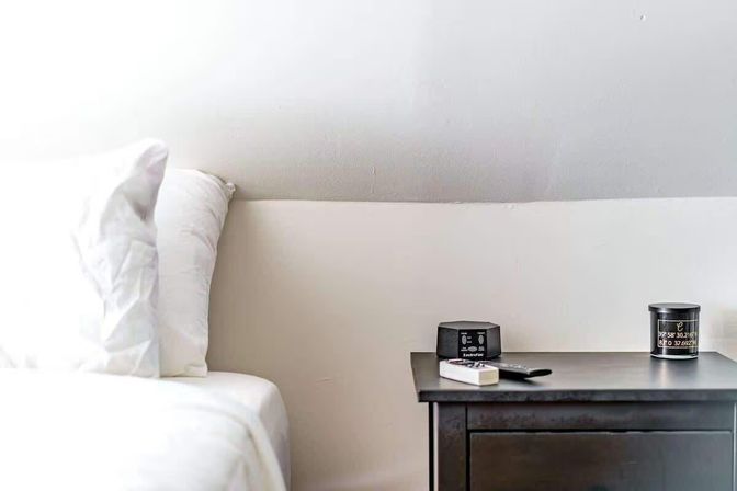 Cozy minimalist bedroom corner with white pillows and sheets beside a dark wood nightstand holding a digital alarm clock, TV remote, and small black candle against a pale wall.