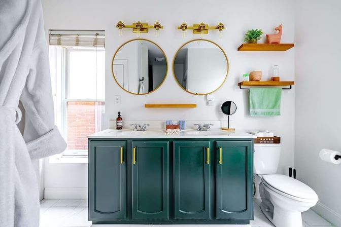 Bright, airy modern bathroom with a deep green double-sink vanity and brass hardware, twin gold-rimmed round mirrors, wooden floating shelves, toilet, and a window.