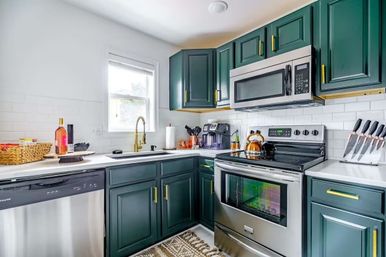 Sunlit modern kitchen with deep green shaker cabinets, white subway tile backsplash, gold hardware and faucet, stainless steel stove, microwave and dishwasher, white countertops and patterned rug