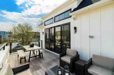 Sunny rooftop deck of a modern white home with black-framed sliding glass doors, gray wood decking, bench-style picnic table, cushioned lounge chairs and glass railing overlooking a neighborhood.