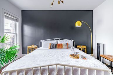 Cozy modern bedroom with white metal bed and textured white bedding against a dark charcoal accent wall, gold arc floor lamp, rattan nightstands, colorful throw pillows, large window and indoor palm plant.
