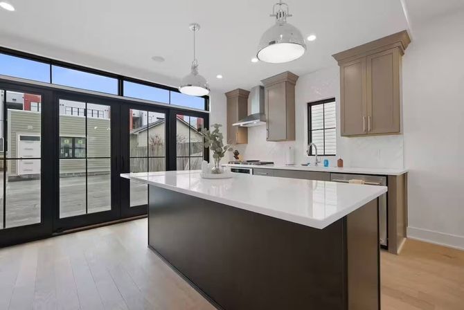Bright modern open-plan kitchen with large white quartz island and dark base, taupe cabinets, stainless range hood, pendant lights, hardwood floors and black-framed floor-to-ceiling glass doors to the yard