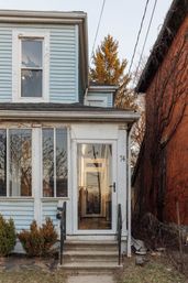 Light-blue two-story house with vinyl siding and glass-enclosed front porch, concrete steps with black railings, narrow brick neighbor and bare trees reflecting sunset light.