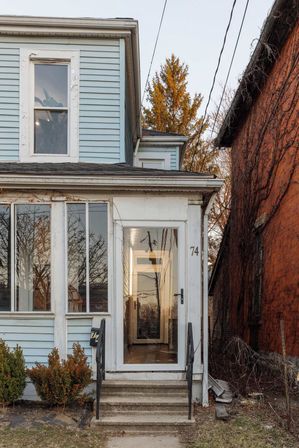 Light-blue two-story house with vinyl siding and glass-enclosed front porch, concrete steps with black railings, narrow brick neighbor and bare trees reflecting sunset light.