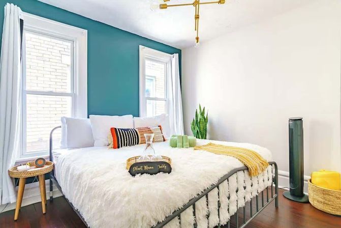 Cozy sunlit modern bedroom with a teal accent wall, fluffy white bedspread, colorful striped pillows and mustard throw, wooden side table, snake plant, brass ceiling light, tall black fan and woven basket on dark hardwood floor.