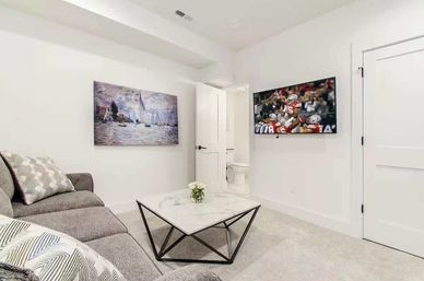 Bright modern living room with gray sectional sofa, geometric marble coffee table, sailboat wall art, wall-mounted TV showing a football game, and an open door to a bathroom.