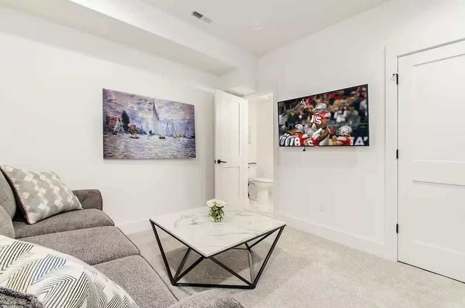 Bright modern living room with gray sectional sofa, geometric marble coffee table, sailboat wall art, wall-mounted TV showing a football game, and an open door to a bathroom.