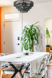 Cozy, plant-filled modern dining room with wooden floors, white-fringed tablecloth on a farmhouse-style table, potted succulent centerpiece, metal chairs, tall indoor plant by the entry door, and a tiered black woven chandelier overhead.