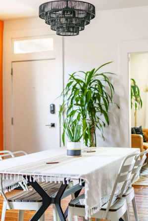 Cozy, plant-filled modern dining room with wooden floors, white-fringed tablecloth on a farmhouse-style table, potted succulent centerpiece, metal chairs, tall indoor plant by the entry door, and a tiered black woven chandelier overhead.