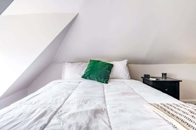 Cozy minimalist attic bedroom with sloped ceiling, white comforter, green accent pillow and black nightstand holding a book and cup