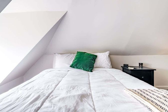 Cozy minimalist attic bedroom with sloped ceiling, white comforter, green accent pillow and black nightstand holding a book and cup