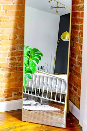 Full-length mirror against exposed brick, reflecting a sunlit loft bedroom with a white metal bed frame, large green monstera plant, gold floor lamp and hardwood floors.
