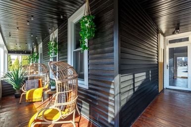 Sunlit covered front porch with dark horizontal siding, two rattan egg chairs with yellow throws, hanging greenery, potted plants, wood floor and string lights.