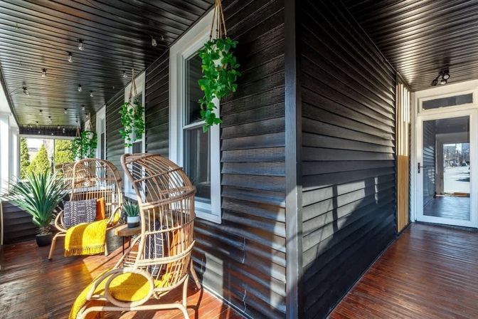 Sunlit covered front porch with dark horizontal siding, two rattan egg chairs with yellow throws, hanging greenery, potted plants, wood floor and string lights.