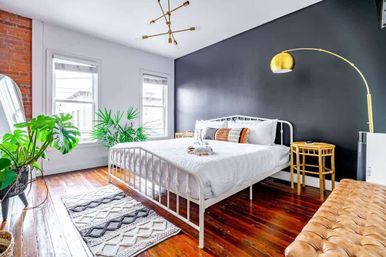 Bright modern bedroom with a black accent wall, white metal bed dressed in white linens and patterned pillows, warm hardwood floors, large green houseplants, brass arc floor lamp and woven rug.