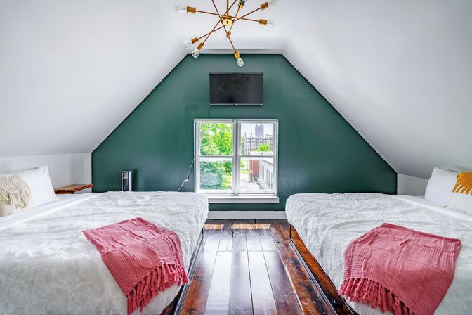 Cozy attic loft bedroom with two beds in white faux-fur bedding and pink throws, dark hardwood floors, green feature wall with wall-mounted TV, modern brass chandelier, and a window showing urban greenery and a distant skyline.
