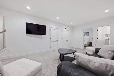 Cozy modern finished basement living room with wall-mounted flat-screen TV, gray carpet, recessed ceiling lights, neutral upholstered seating and a black triangular coffee table.