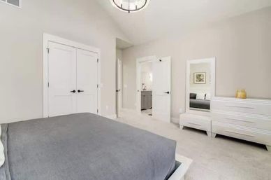 Bright modern master bedroom with gray bedding, white double-door closet, ensuite bathroom, white dresser and full-length mirror on a carpeted floor.