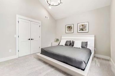 Modern minimalist bedroom interior with white platform bed and gray bedding, four pillows, nightstand with flowers, two abstract wall prints above the headboard, white double closet doors, vaulted ceiling with round pendant light, soft gray carpet and neutral walls.
