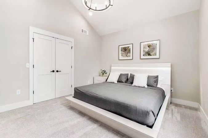 Modern minimalist bedroom interior with white platform bed and gray bedding, four pillows, nightstand with flowers, two abstract wall prints above the headboard, white double closet doors, vaulted ceiling with round pendant light, soft gray carpet and neutral walls.
