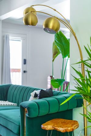 Bright modern living room with emerald green velvet channel-tufted sofa, gold arc floor lamp with three globe shades, lush tropical houseplants and a live-edge wooden side table
