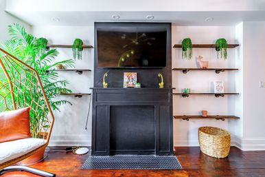 Bright modern living room with black fireplace mantel and wall-mounted TV, wooden floating shelves with trailing green plants, rattan hanging chair with leather cushion, large potted palm and woven basket on hardwood floors.