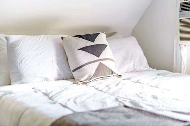 Bright minimalist loft bedroom with crisp white comforter, geometric black-and-white accent pillow and a window air conditioner