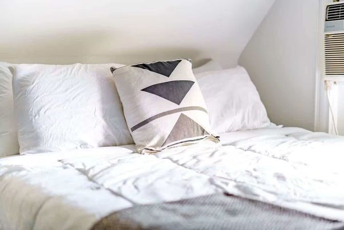 Bright minimalist loft bedroom with crisp white comforter, geometric black-and-white accent pillow and a window air conditioner